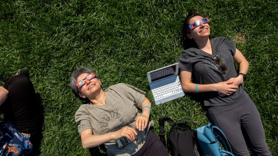 Two women, with a laptop between them, sitting on grass and looking up a soalr eclipse while wearing safety sunglasses.