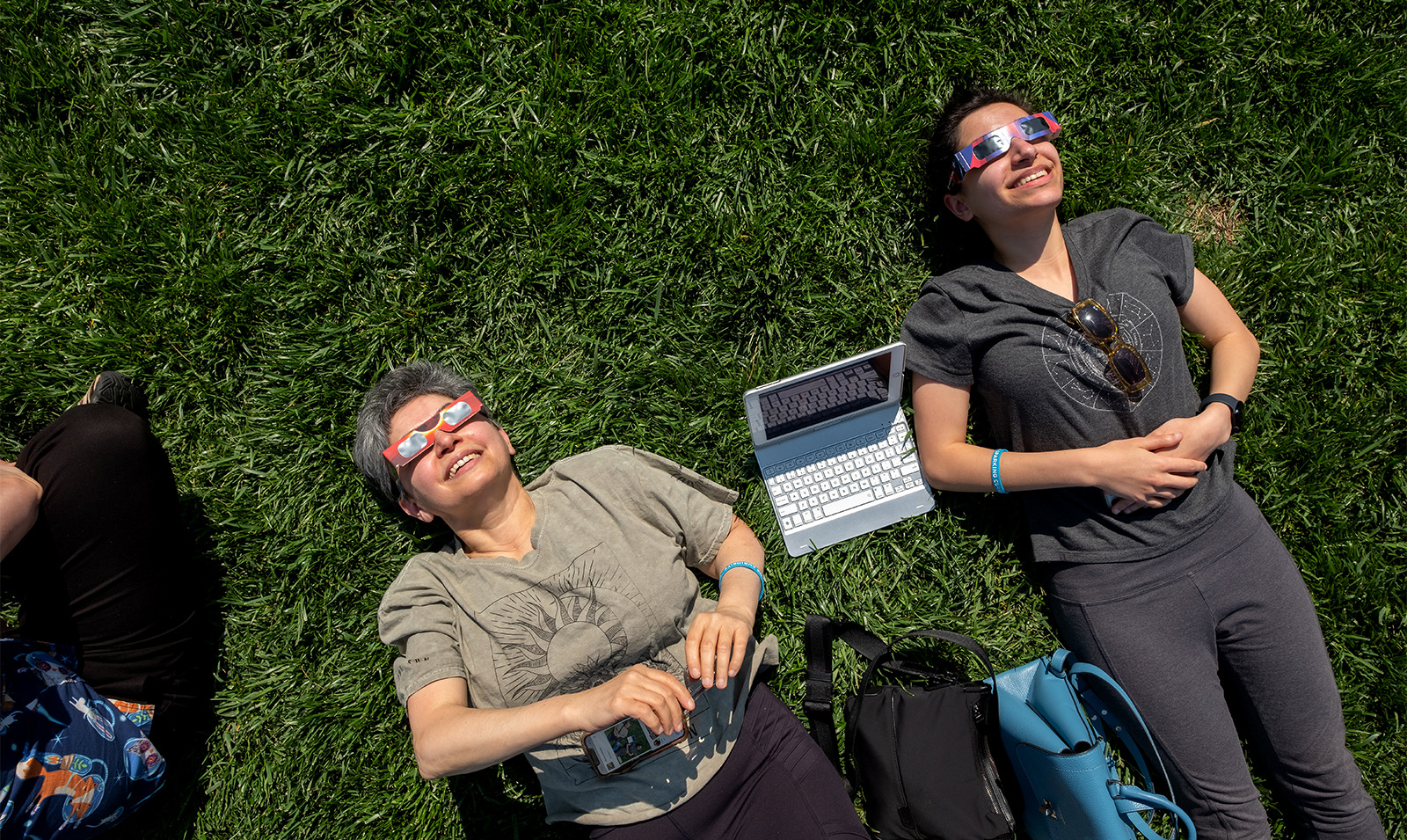 Two women, with a laptop between them, sitting on grass and looking up a soalr eclipse while wearing safety sunglasses.
