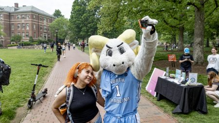 A ram mascot named Rameses taking a selfie with a student on a brick pathway on the campus of UNC-Chapel Hill.