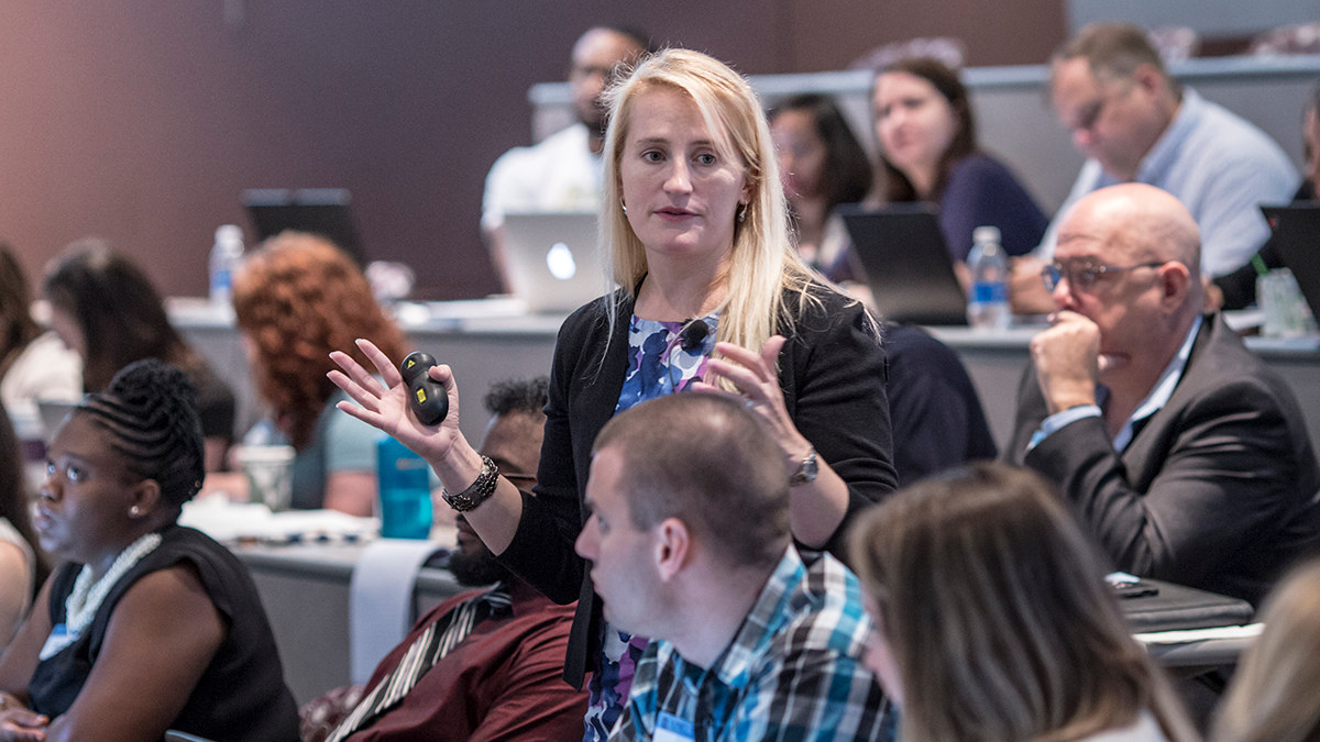 Woman standing up amongst crowd of seated individuals in a lecture hall.