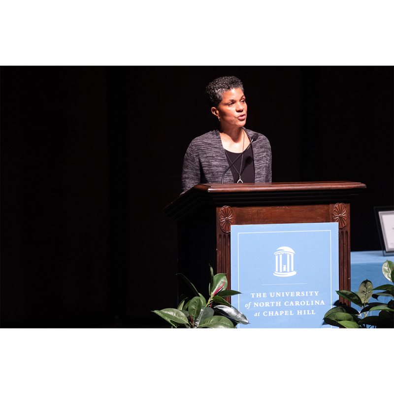A woman, Michelle Alexander, speaking from a podium on a stage at a lecture.