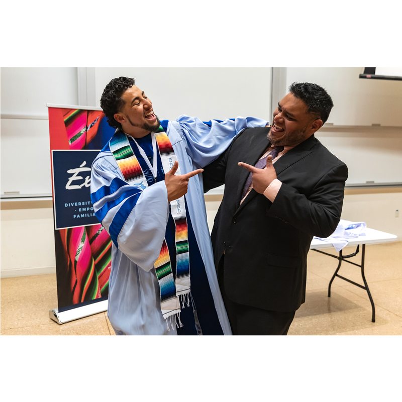 Student in graduation gown and Exitos sash smiles with staff member