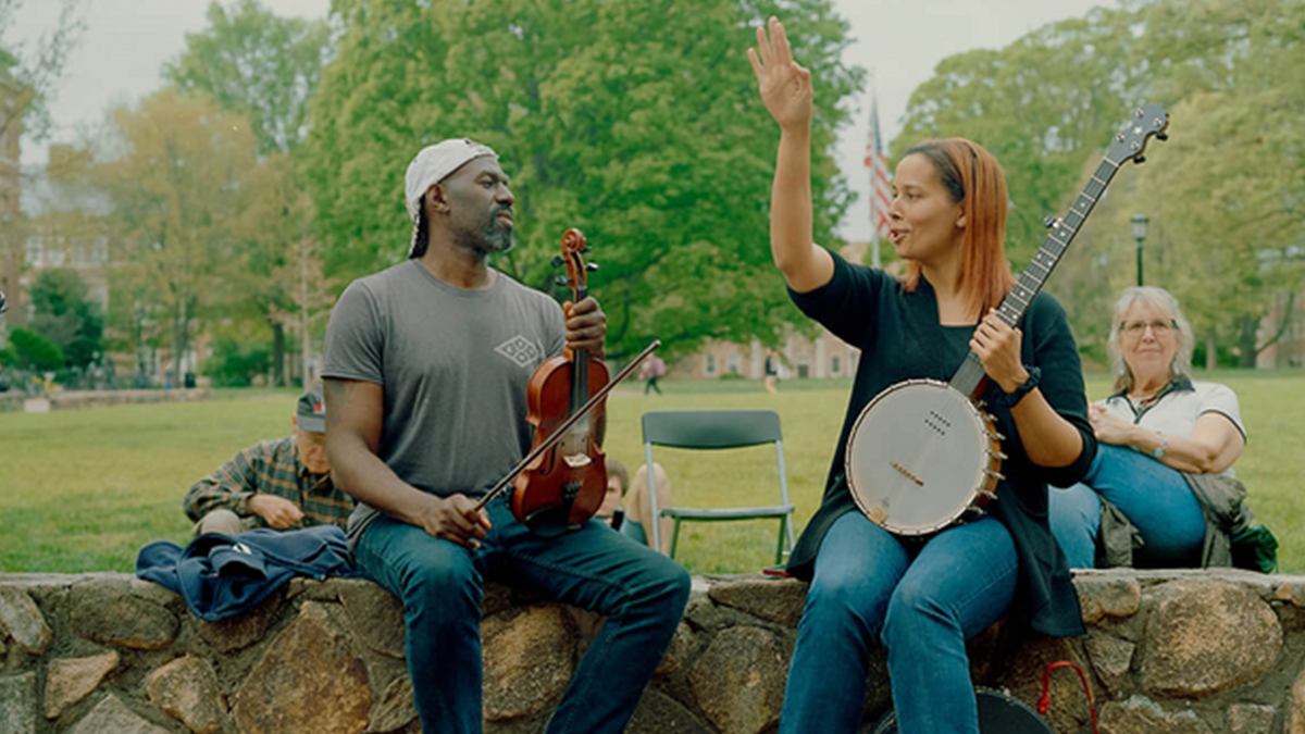 Rhiannon Giddens with banjo in hand and arm outstretched talking to Justin Robinson wearing a baseball camp and holding a violin on the main UNC quad.