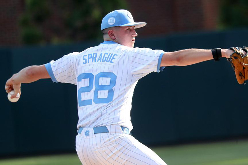 A Carolina baseball player, Shea Sprague, readying to throw a pitch. On his Carolina Blue hat is a patch with the initials "JR."