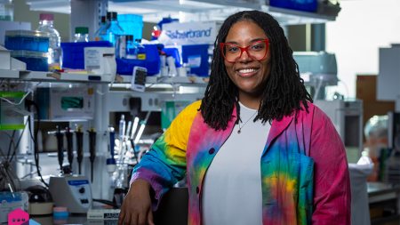 Jasmine King wearing a white shirt and rainbow-colored blazer in front of research equipment.