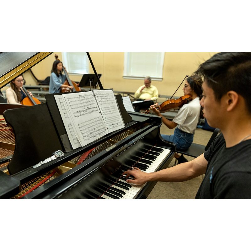 Students in a music class playing a variety of instruments. In the foreground is a student playing the piano and reading sheet music.