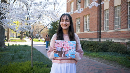 A student, Daneen Khan, posing with a painting she made while standing on a brick pathway next to a building on the campus of UNC-Chapel Hill.