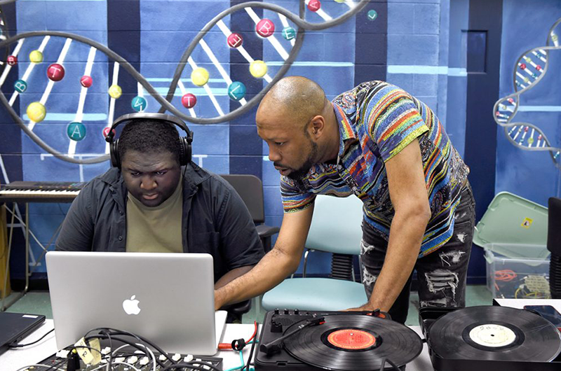 A male instructor leans over a male student wearing headphones at a laptop.