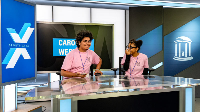 Two high school students sitting at anchor desk of a TV studio on the campus of UNC Chapel Hill.