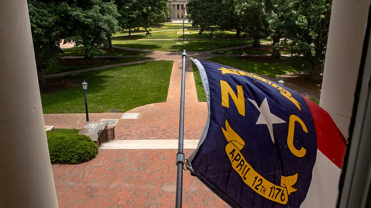 Photograph of the North Carolina flag blowing in the wind - overlooking a quad at UNC.