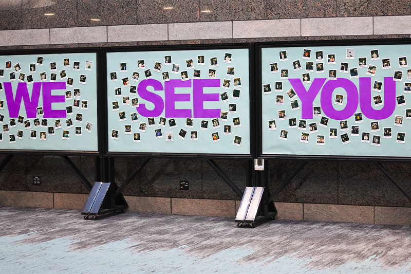 Artwork spread across three bulletin boards reading in large purple letters "WE SEE YOU." In the background of the letters are scattered Polaroid photos of people.