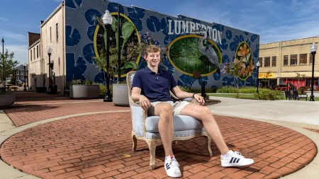 Matthew Foil sitting in a Carolina Blue Chair in downtown Lumberton, with a large building-side 