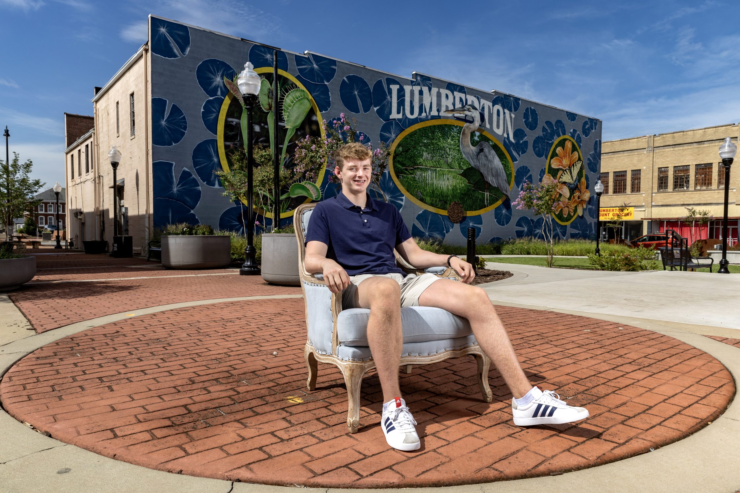 Matthew Foil sitting in a Carolina Blue Chair in downtown Lumberton, with a large building-side 