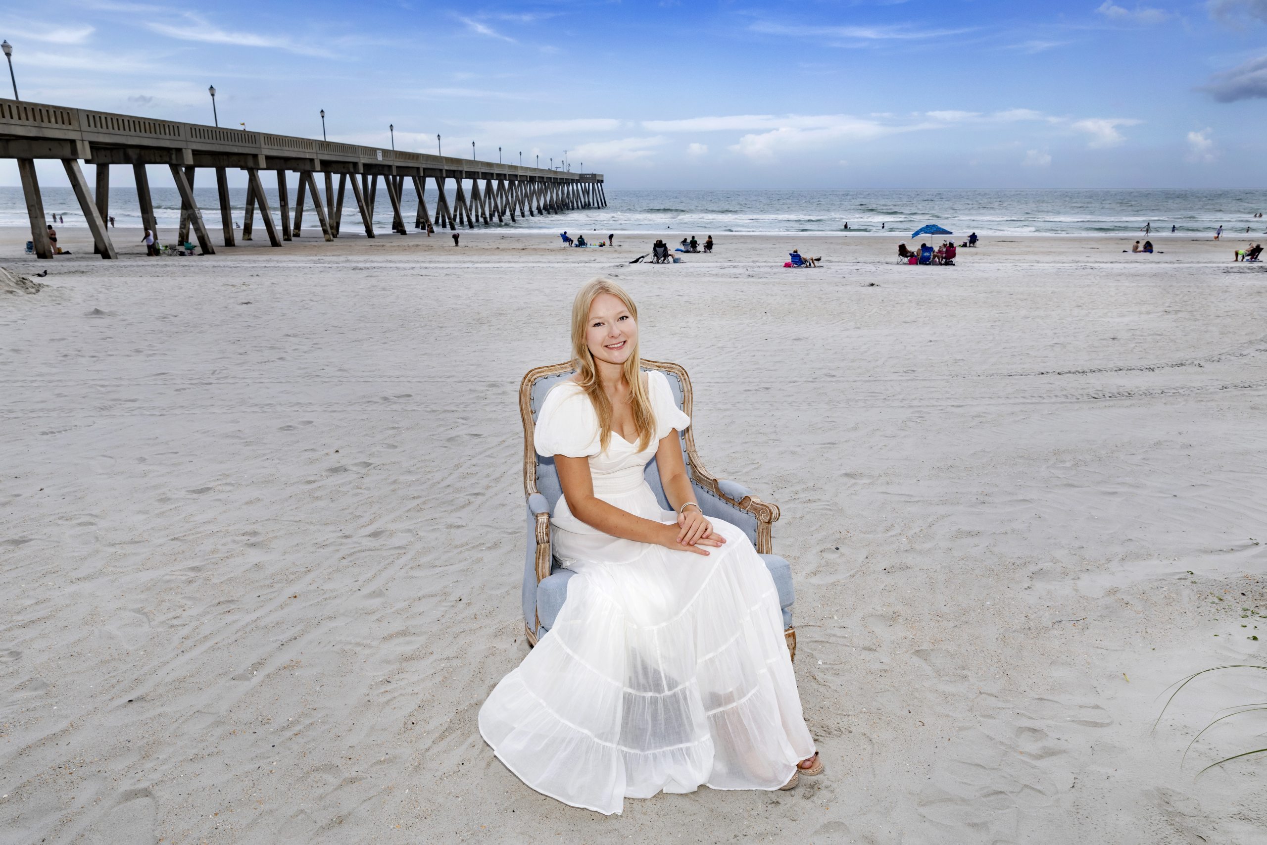 Isabella Cox sitting in a Carolina Blue chair on a beach with the Atlantic Ocean and a pier seen in the background.