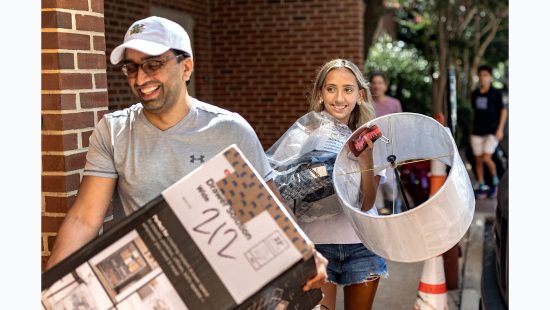 A father and daughter carrying supplies outside of a dormitory building.