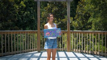 Alyssa Crane outdoors, with trees seen in background, standing and holding one of her paintings.