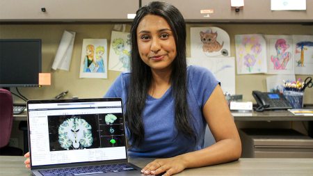 Ria Patel sitting and smiling at a table with an x-ray of a brain on a laptop screen next to her.