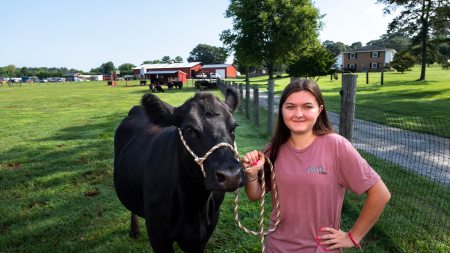 A woman, Rylea Suddreth, posing for a portrait with a Black cow, named Hattie, on grassland on her family farm.