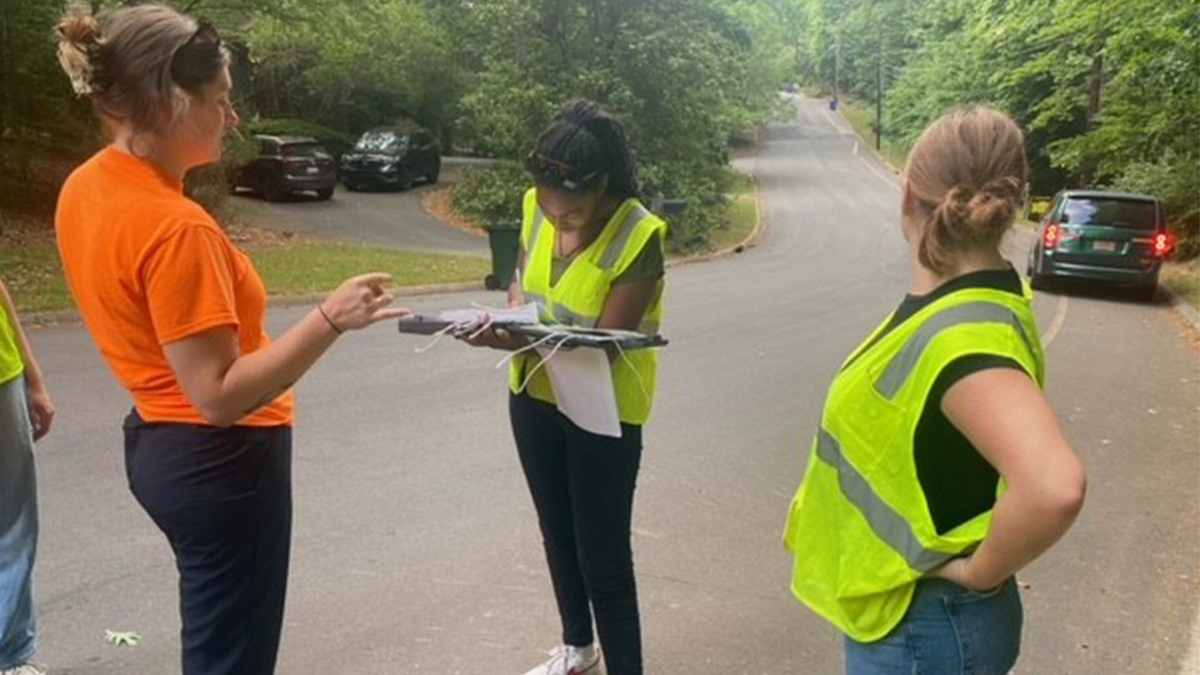 Brianna wearing a yellow vest and holding a clipboard while speaking with citizens on a gravel road.