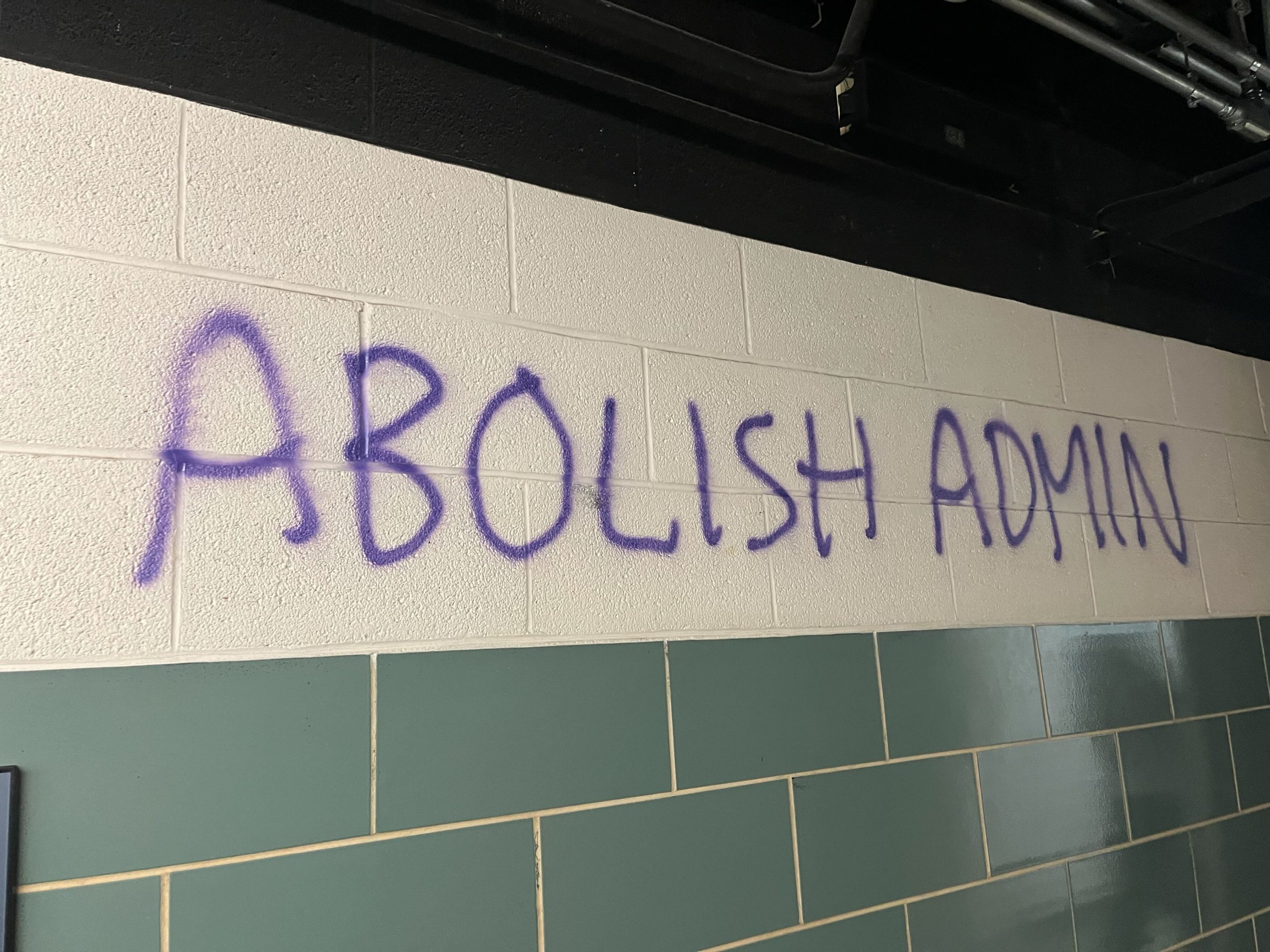 Wall with "ABOLISH ADMIN" spray painted in purple lettering