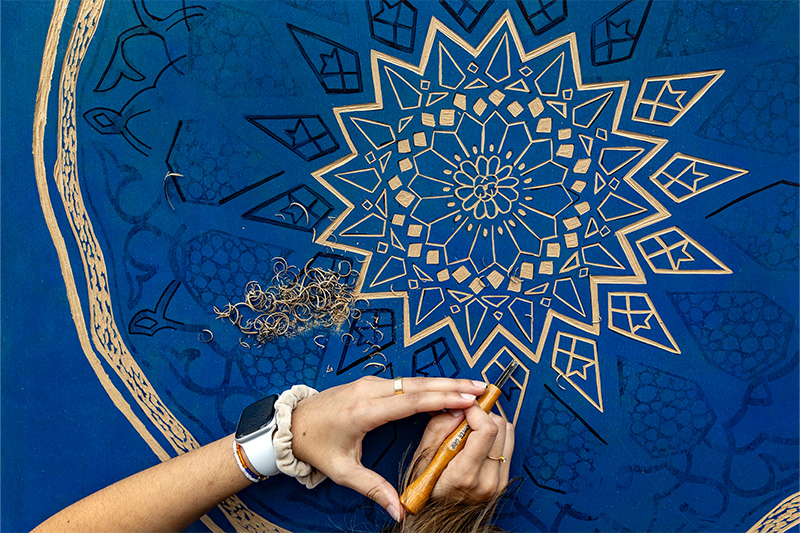 Photo with an angle from above featuring an art piece being worked on by student Aliha Younus, whose hands are seen using a tool to carve on her tapestry.