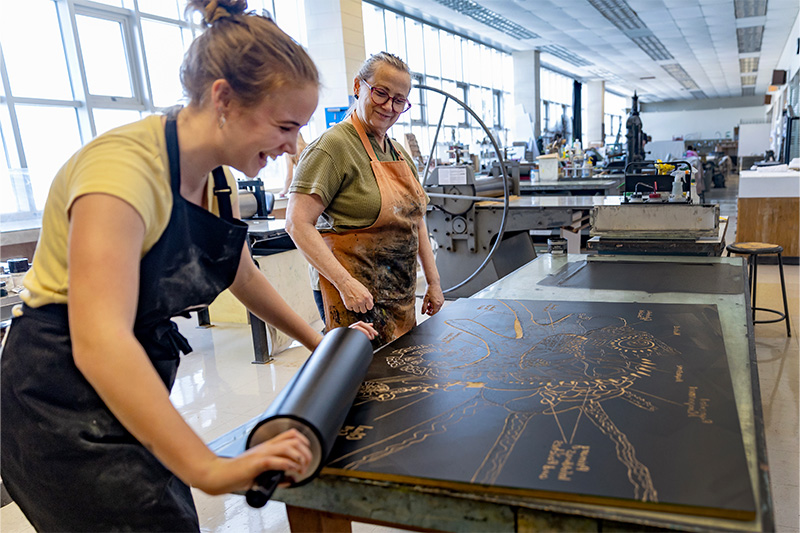 A student, Jacqueline Ari, laughing with a professor, Beth Grabowski, as she uses a rolling tool on an art piece 