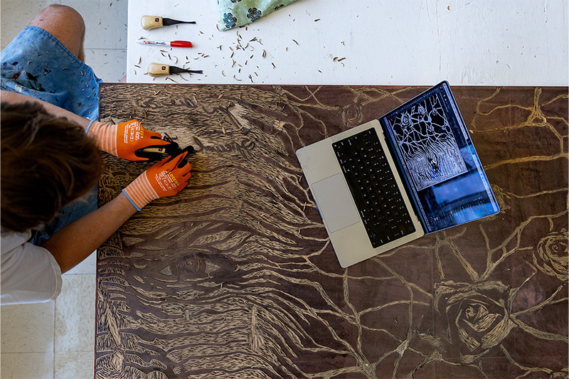 Angle from above of a student using a tool to carve a piece of a large tapestry they're working on with the student's laptop also in sight.