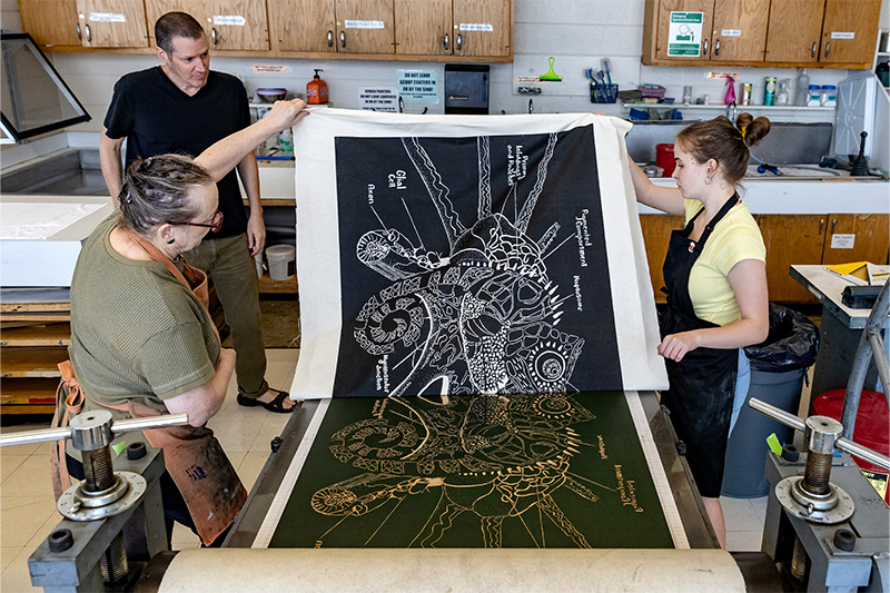 Professors Bob Goldstein and Beth Grabowski examining a large tapestry of art created by student Jacqueline Ari.