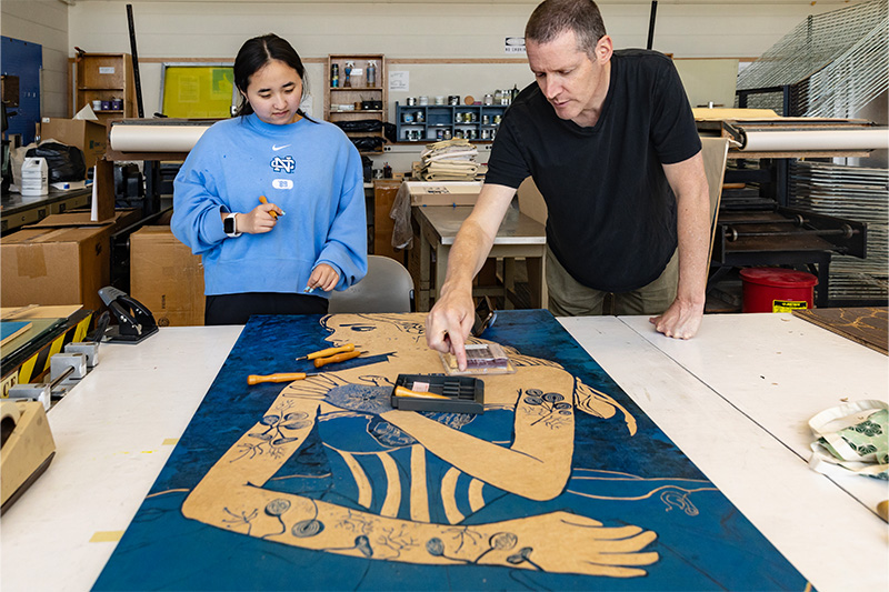 A student, Sophia Oh, and a professor, Bob Goldstein, looking a large painting Oh is working of. The painting is of a woman with a dark blue background.