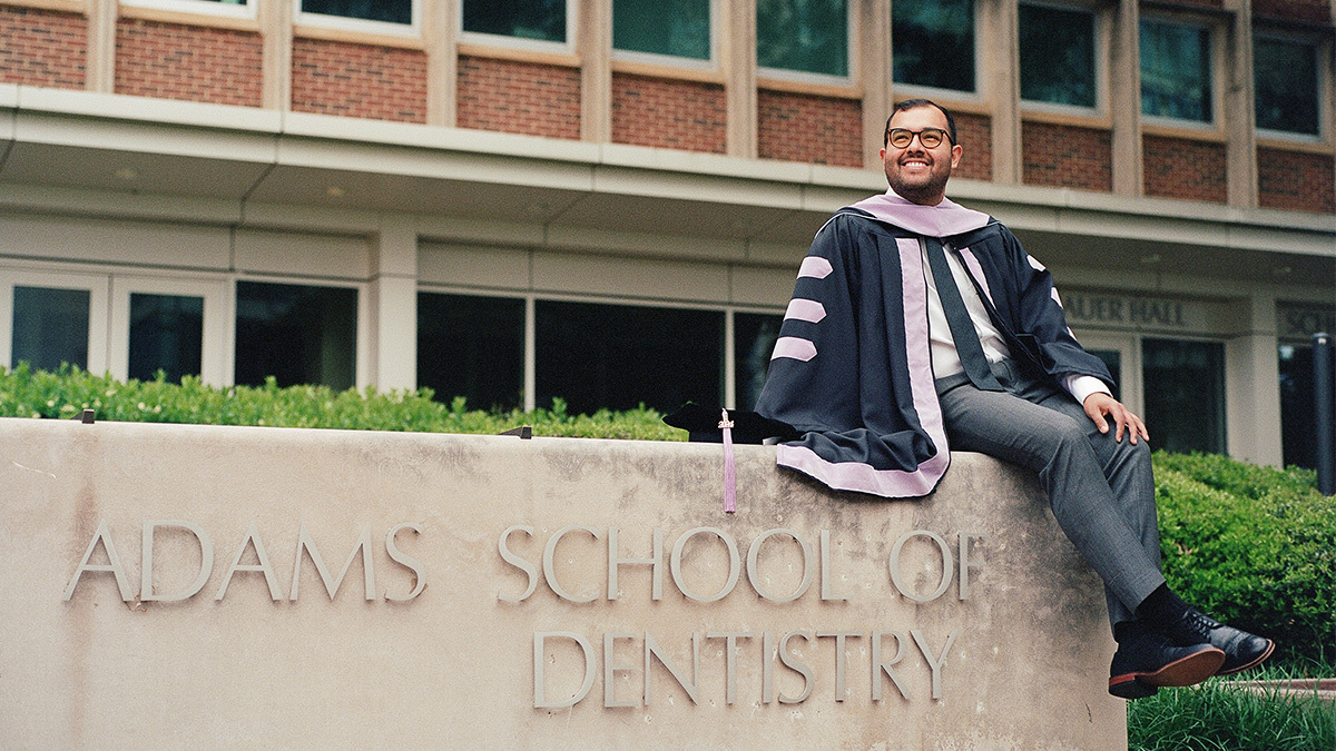 Bruno Segovia-Chumbez wearing commencement robes and sitting on wall reading 