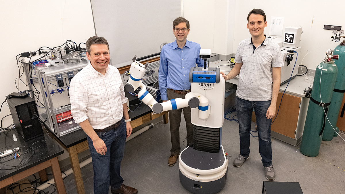 Three graduate students pose for a photo around the robotic arm.