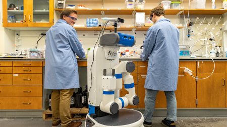 Two chemistry graduate students wearing in lab coats work at a lab bench while an automaton robot with a single, long arm prepares to work near them.