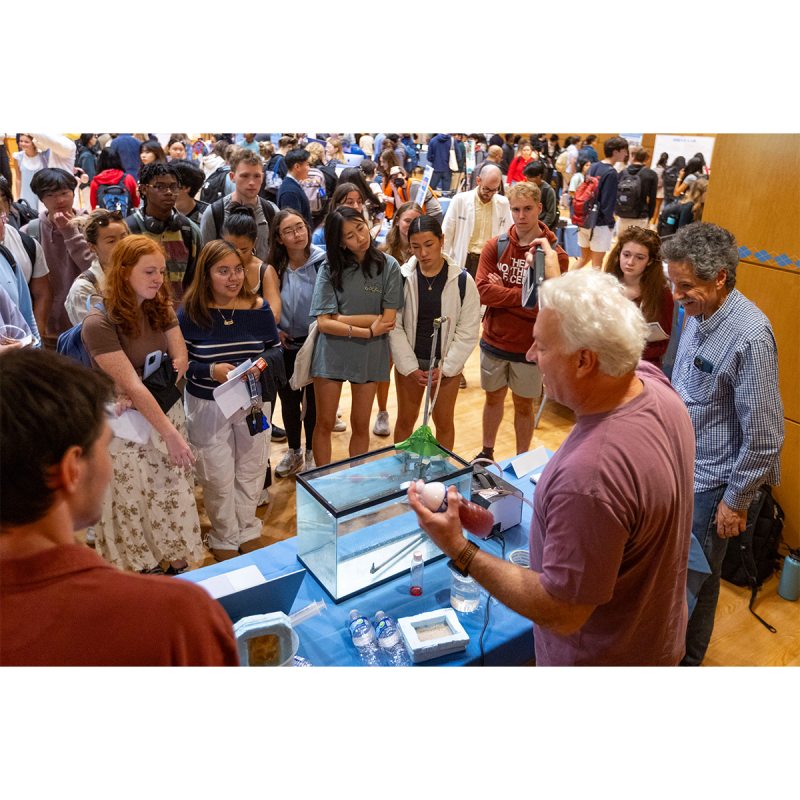 Professors talking to a group of students at a Research Week fair on the campus of UNC-Chapel Hill inside of a hall.