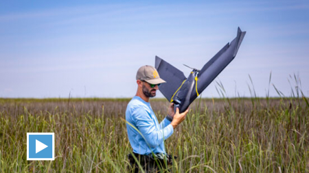 A man carries a drone out of a marsh.