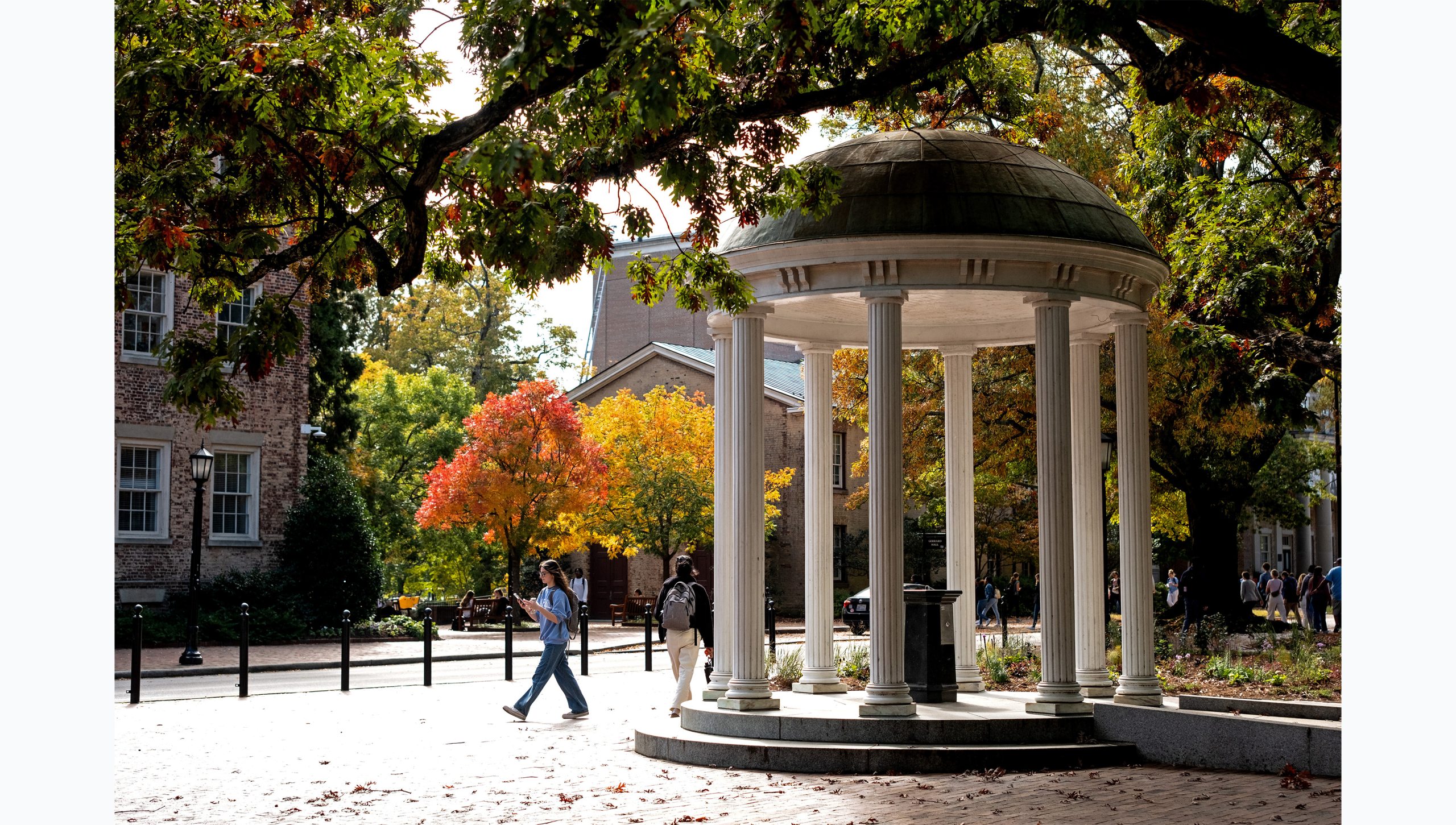 A student walks by the Old Well on the campus of UNC-Chapel Hill. Trees with red, yellow and green leaves are seen in the background.