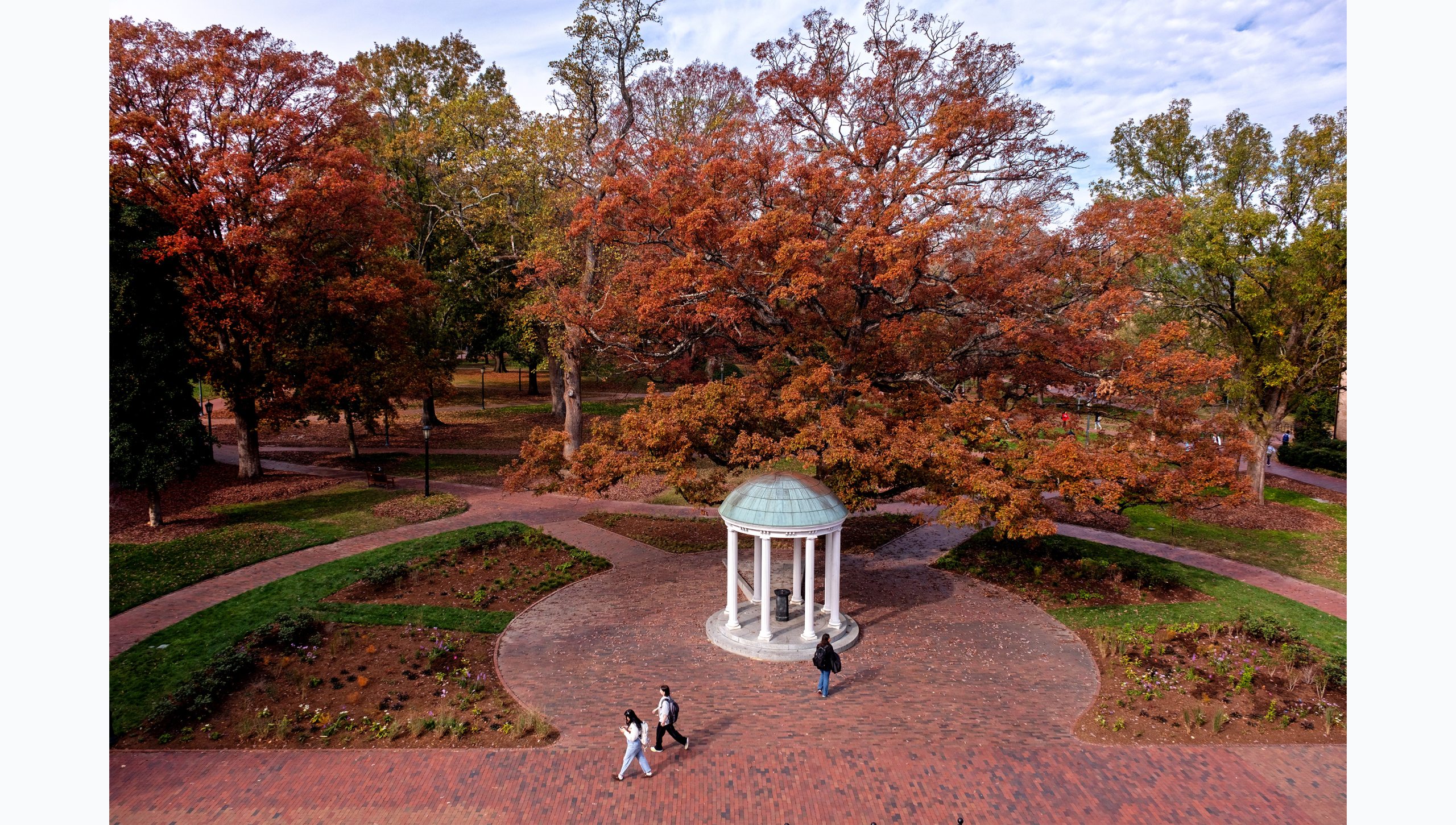 Aerial view of the Old Well on the campus of UNC-Chapel Hill on a fall afternoon. Students are seen walking by on brick pathways, and trees with orange, red, yellow and green leaves are pictured in the background.