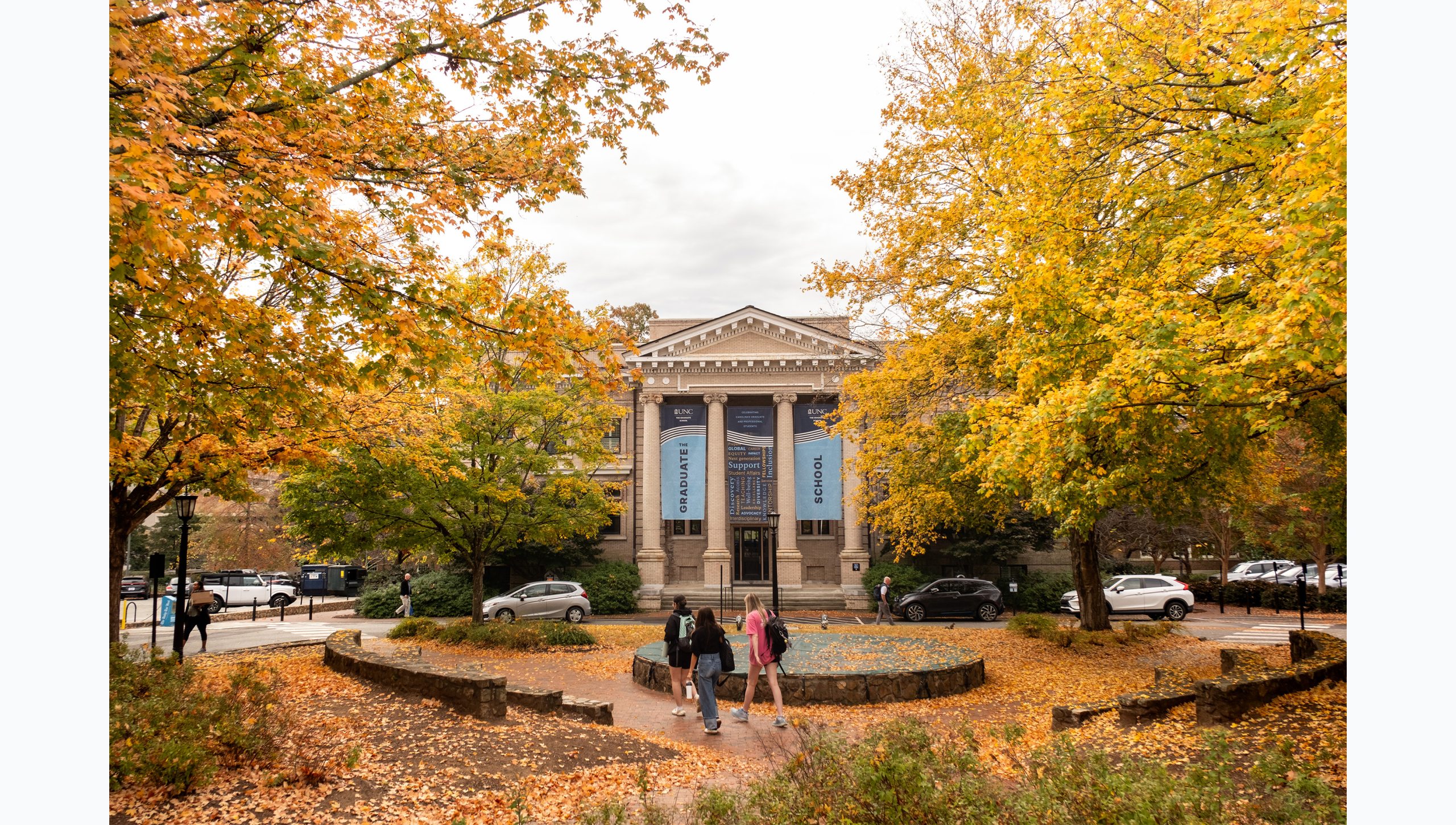 Students walking by the fountain in front of Bynum Hall on the campus of UNC-Chapel Hill. Trees with yellow and green leaves are seen in the background.