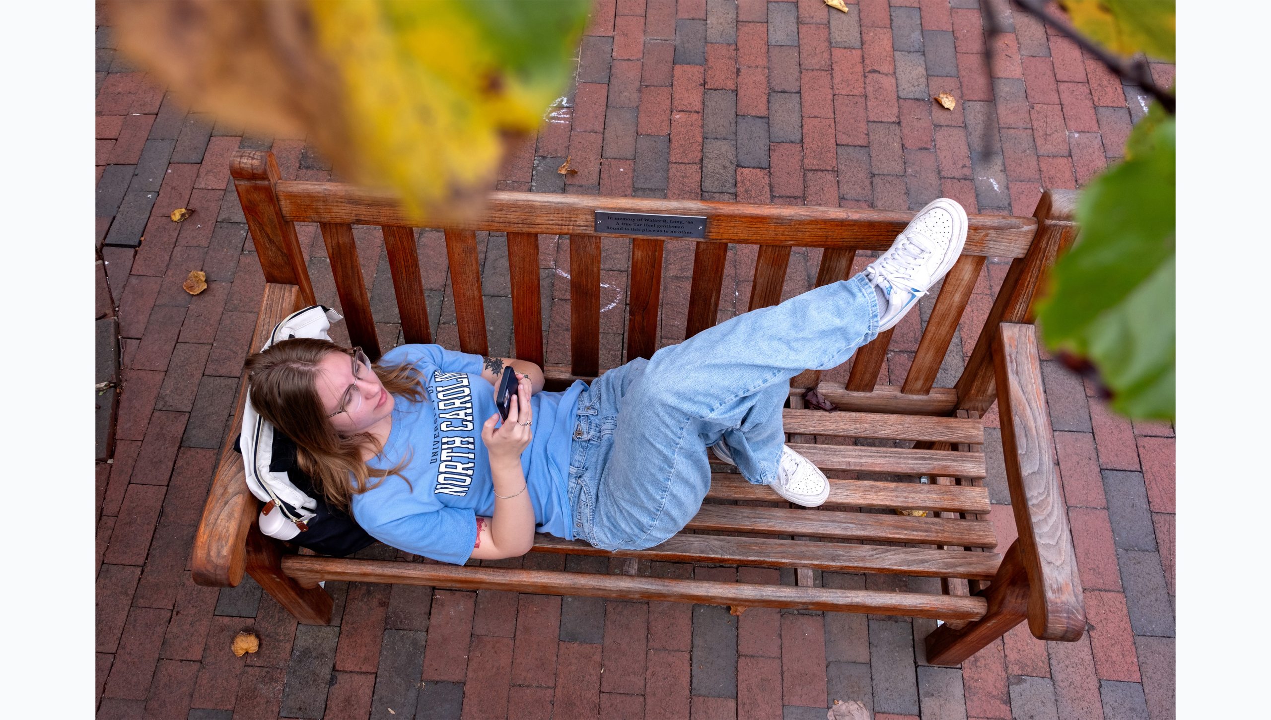 Overhead angle of a student sitting on a wooden bench outside on the campus of UNC-Chapel Hill. Bits of leaves of an overhanging tree are seen as well.