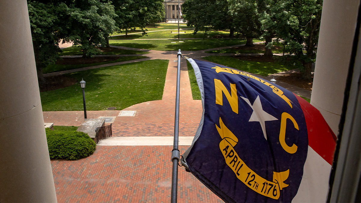 North Carolina state flag flying outside South Building on the campus of UNC-Chapel Hill with Polk Place seen in the background.