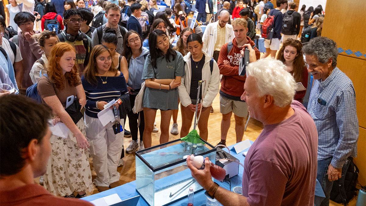 Group of UNC-Chapel Hill students at a research fair attending a demonstration by two professors who conduct fluid research.