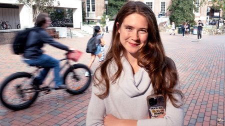 Keegan Lee posing for a portrait on brick pathways on the campus of UNC-Chapel Hill. She's holding her phone, which has a sticker on it reading: 