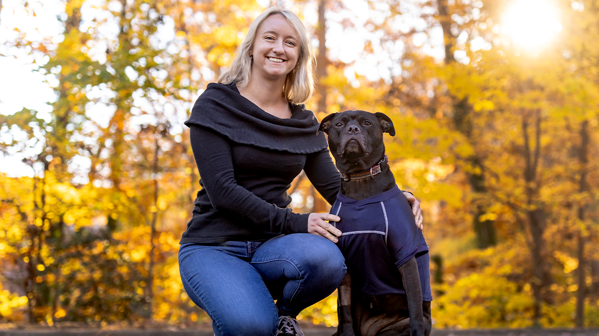 Caitlin Russell posing with her dog, Lumen against autumnal backdrop.